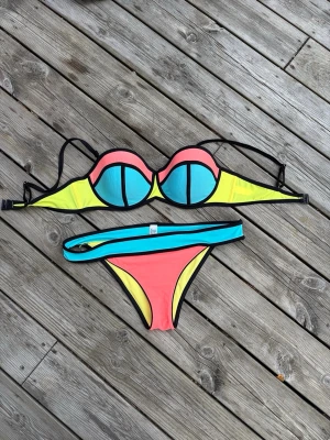 Färgglad bikini med svarta kanter - Skriv gärna innan köp!!! Säljer en bikini i färgglada block med turkos, gul och korallrosa. Överdelen har bygel och vadderade kupor med svarta kontrastkanter och justerbara axelband. Underdelen har matchande färgblock och svart kant. Materialet är stretchigt och snabbtorkande.