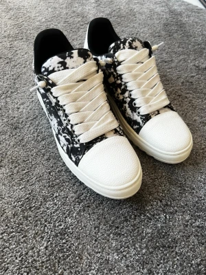 Svartvita sneakers med mönster - Snygga sneakers i svart och vitt med ett unikt marmorerat mönster. Skorna har vita, breda skosnören och vit tå samt sula. Låg modell med räfflad yttersula och svart vadderad häl. Perfekta för dig som vill sticka ut med din stil.