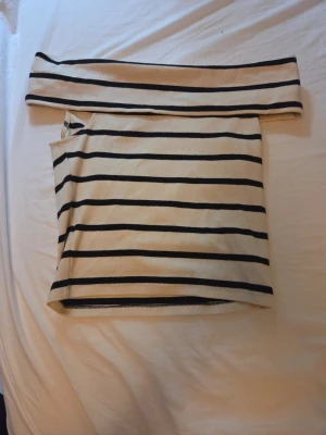 Randig offshoulder topp Gina Tricot - Säljer en snygg offshoulder topp från Gina Tricot i vitt med svarta horisontella ränder. Toppen är axelbandslös och har en tight passform, perfekt för dig som gillar en trendig och stilren look. Materialet är mjukt och stretchigt, troligen bomull eller en bomullsblandning.