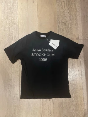Svart Acne Studios t-shirt Stockholm - Snygg svart t-shirt från Acne Studios med trycket 'Acne Studios STOCKHOLM 1996' på bröstet. Klassisk passform med korta ärmar och rund hals. Tillverkad i mjuk bomull för en skön känsla. Perfekt för dig som gillar stilrena och trendiga plagg. Modellen är 182 och 65 kg