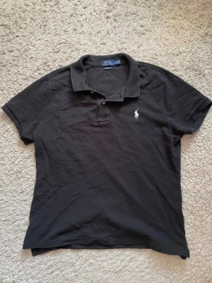 Svart pikétröja från Polo Ralph Lauren - Klassisk svart pikétröja från Polo Ralph Lauren med vit broderad logga på bröstet. Tröjan har korta ärmar, krage och knappar framtill. Tillverkad i mjuk bomull som känns skön mot huden. Perfekt till jeans eller shorts. Kolla på alla bilder noga❗️den sitter som en tajt M ❗️på bilderna ser den brun/röd ut, detta är bara på bilderna och ljuset❗️