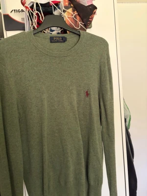 Grön stickad tröja från Polo Ralph Lauren - Snygg grön stickad tröja från Polo Ralph Lauren med klassisk rund halsringning och den ikoniska röda logotypen broderad på bröstet. Tröjan har lång ärm och är tillverkad i mjukt material som passar perfekt till alla tillfällen.