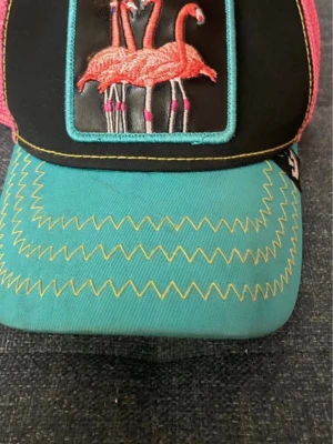 Turkos keps med flamingos och text - Snygg keps med turkos skärm och svart frontpanel. På framsidan finns ett broderat märke med fyra rosa flamingos och texten 'SQUAD' i gult. Skärmen har dekorativa gula sicksacksömmar och kepsen har mesh på sidorna. Perfekt för dig som gillar färg och unika detaljer.