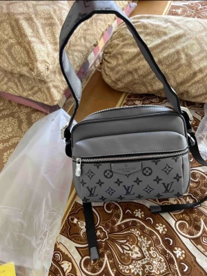 Grå axelväska  - Snygg grå axelväska med klassiskt monogrammönster i svart. Väskan har justerbar axelrem, två dragkedjefack och är tillverkad i ett tåligt material med stilren design. Perfekt för dig som vill ha en trendig och praktisk väska.
