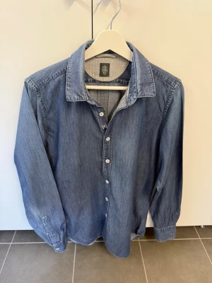 Blå Eleventy skjorta, stl 40/M - Snygg blå jeansskjorta med klassisk krage och vita knappar. Skjortan har lång ärm och är tillverkad i ett mjukt bomullsmaterial. Perfekt för en avslappnad och trendig look.