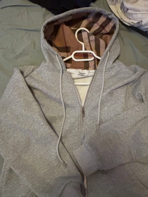 Grå zip hoodie från Burberry - Snygg grå zip hoodie från Burberry med klassiskt rutigt foder i huvan. Hoodien har dragsko i huvan, ribbade muddar och är tillverkad i mjukt material. Perfekt för en chill och stilren look. Tröjan passar har storleken small/S men passar bäst som medium/M.