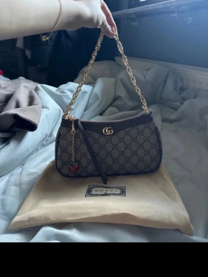 Gucci GG Supreme axelremsväska - Snygg handväska från Gucci i GG Supreme-mönstrad canvas med bruna läderdetaljer och guldfärgad kedjerem. Väskan har ett klassiskt G-logga framtill och ett litet jordgubbshänge. Perfekt storlek för det viktigaste och ger en lyxig touch till din outfit.