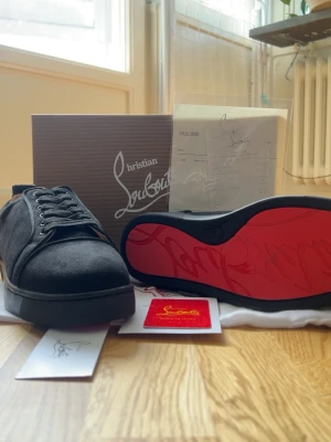 Svarta sneakers från Christian Louboutin - Snygga svarta sneakers från Christian Louboutin med klassisk röd sula och svarta skosnören. Skorna har en stilren design i mocka och låg profil som passar till många olika outfits. Kommer med originalkartong och tillbehör.