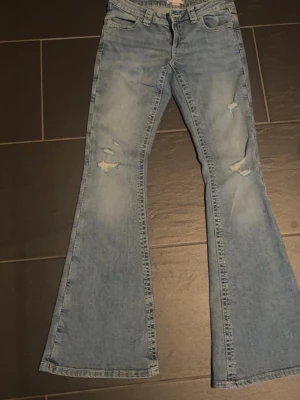 Ljusblå utsvängda jeans med slitningar - Säljer ett par ljusblå jeans med utsvängda ben och slitningar på framsidan. Jeansen har klassisk femficksmodell och broderade detaljer på bakfickorna. Perfekta för dig som gillar retrostil och vill sticka ut lite extra.   Jeans ifrån gina 14+ aldrig använda. Storlek S