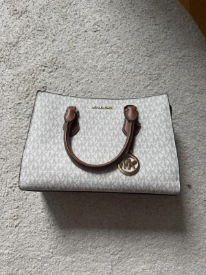 Vit handväska från Michael Kors - Snygg vit handväska från Michael Kors med bruna handtag och guldfärgade detaljer. Väskan har MK-mönster över hela ytan, ett hängande MK-emblem och flera fack med dragkedja. Perfekt för dig som gillar stilrena accessoarer.