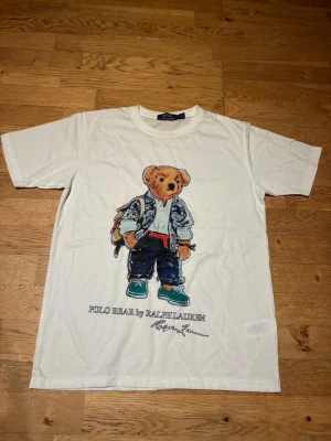Vit Polo Bear t-shirt från Ralph Lauren - Vit t-shirt från Ralph Lauren med ikoniskt Polo Bear-tryck framtill. T-shirten har rund hals och korta ärmar. Tillverkad i mjuk bomull och har en avslappnad passform. Perfekt för dig som gillar streetwear och klassiska prints.                                     Kontakta vid frågor och prisförslag. Skickar inom 24h.