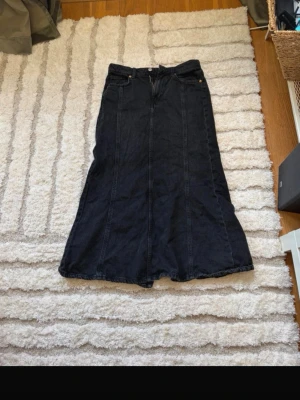 Svart lång jeanskjol - Säljer en svart lång jeanskjol med klassisk femficksdesign och synliga sömmar. Kjolen har en lätt utsvängd form och stängs med knapp och dragkedja framtill. Perfekt för dig som gillar en enkel men snygg stil. Även slits där bak 