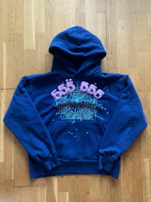 Blå Sp5der hoodie  - Blå hoodie med huva och känguruficka. På bröstet finns ett stort tryck av ett grönt spindelnät med små stjärnor och siffrorna '555' i rosa 3D-tryck ovanför. Hoodien har ribbade muddar och en avslappnad passform. Perfekt för dig som gillar unika prints.