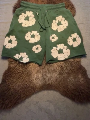 Denim Tears shorts - Gröna shorts med vita blommiga tryck och snörning i midjan. Shortsen har elastisk midja och en bakficka. Perfekta för sommarens alla äventyr och har en avslappnad passform.