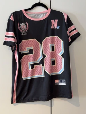 Svart t-shirt med rosa detaljer och tryck - Cool svart t-shirt med stora siffran 28 i rosa och vitt framtill, rosa och vita ränder på ärmarna samt sportiga tryck på bröstet. T-shirten har rund halsringning och korta ärmar. Perfekt för en avslappnad och sportig look.