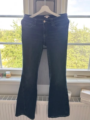 Mörkblå bootcut jeans med broderi - Snygga mörkblå bootcut jeans med kontrastsömmar och broderade detaljer på bakfickorna. Jeansen har klassisk femficksmodell och vita knappar på fickorna bak. Perfekta för dig som gillar en retroinspirerad look.