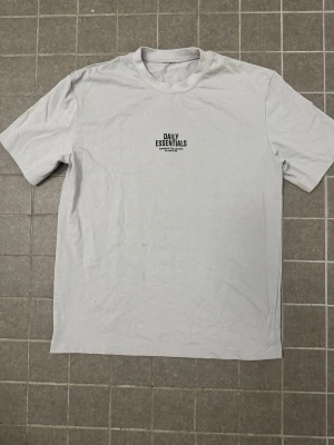  Essentials t-shirt - Snygg grå oversized t-shirt med tryck från Daily Essentials. Framsidan har en liten logga på bröstet och baksidan har ett stort tryck med texten 'DAILY ESSENTIALS' och detaljer i svart. Jätte bra och billig pris/skick!