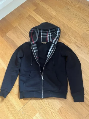 Burberry Tröja - Snygg svart hoodie med dragkedja och klassiskt rutigt foder i huvan. Hoodien har två fickor framtill, justerbara snören i huvan och ribbade muddar vid ärmar och midja. Perfekt för en chill och stilren look.
