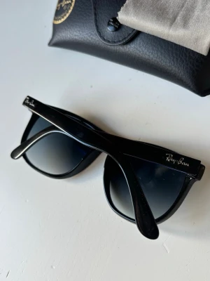 Svarta Ray-Ban Wayfarer solglasögon - Klassiska svarta Ray-Ban Wayfarer solglasögon med snyggt, tidlöst utseende. Bågarna är i blank svart plast och glasen är mörka med en lätt toning. Perfekta för att höja din stil och skydda ögonen mot solen. Medföljer originalfodral och putsduk.