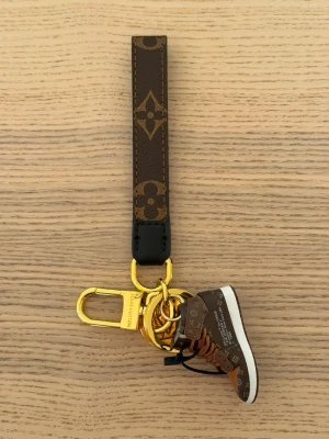  Vuitton nyckelring med sneaker - Unik nyckelring från  Vuitton med klassiskt monogrammönster i brunt och guld. Nyckelringen har en miniatyrsneaker i brunt och vitt, guldfärgade metalldetaljer och LV-logga. Perfekt accessoar för att piffa till väskan eller nyckelknippan.