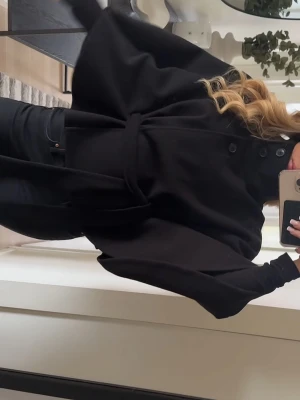 Omlott kappa  - Stilren svart cape-kappa med knytband i midjan och bred krage. Kappan har knappar vid axeln och vida ärmar för en trendig siluett. Perfekt för dig som vill ha en modern och elegant look. Materialet ser ut att vara mjukt tyg, troligen ullblandning.