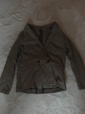 Brunrutigt dubbelknäppt kavaj - Snygg kavaj med brunt och vitt rutmönster och dubbelknäppning framtill. Klassisk krage och långa ärmar ger en stilren look. Perfekt för dig som vill ha en trendig och tidlös stil. Materialet känns mjukt och bekvämt.
