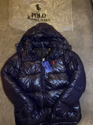 Pufferjacka Polo Ralph Lauren Shiny Navy 🐎 - Brand new Polo Ralph Lauren puffer jacket in a glossy navy blue finish, featuring a cozy hood and the iconic red pony logo on the chest. This stylish and warm jacket comes with original tags attached, indicating pristine condition.  ⭐️ Condition : 10/10 (New with tags) 🎨 Color : Glossy Navy Blue 📦 Fast and careful shipping