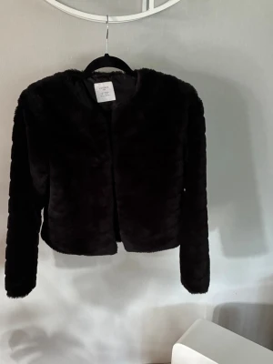 Svart kort pälsjacka - Snygg svart pälsjacka i fuskpäls med rund halsringning och lång ärm. Jackan är kort i modellen och har en stilren, minimalistisk look. Stängs med hyska och hake framtill, tre från början en kvar. Perfekt för att höja din outfit under kalla dagar.