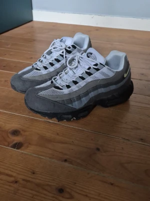 Nike Air Max 95 grå sneakers - Nike Air Max 95 sneakers i olika nyanser av grått med vita meshpaneler och svarta detaljer. Skorna har synlig Air-dämpning i sulan och klassisk snörning. Swoosh-logga på sidan och retroinspirerad design. Perfekta för dig som gillar streetwear och sportig stil. 