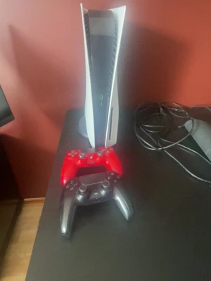 PlayStation 5 med två handkontroller - Säljer en PlayStation 5 konsol i vit färg med futuristisk design. Medföljer två trådlösa handkontroller, en röd och en svart. Perfekt för gaming med vänner och familj. Konsolen har en stilren look och står vertikalt. Ps5 kontrollen knappen R2 är sönder men ps4 funkar perfekt har använt i 1.5 år