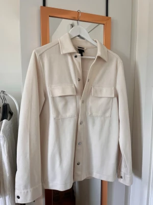 Vit overshirt med fickor - Stilren overshirt med två stora bröstfickor och tryckknappar framtill. Overshirten har klassisk krage och långa ärmar med knapp vid ärmslut. Tillverkad i ett mjukt tyg.