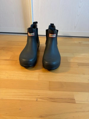 Svarta Chelsea boots från Hunter - Säljer ett par svarta Chelsea boots från Hunter i gummi med klassisk siluett och elastiska paneler på sidorna. Bootsens dragflikar gör dem enkla att ta på och av. Perfekta för regniga dagar och har en stilren look som funkar till många outfits.
