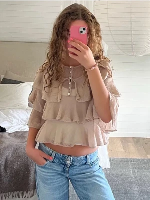 Beige volangblus med knappar - Supersöt beige blus med flera volanglager och knappar framtill. Croppad modell med trekvartsärm och rund halsringning. Supersnygg att styla med ett par basic jeans eller en fin kjol! Från Zara i storlek M.