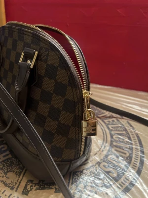 Brun-rutig handväska med gulddetaljer - Säljer en snygg handväska i brun och mörkbrun rutig design med guldfärgad dragkedja och lås. Väskan har både handtag och axelrem samt röd insida. Perfekt för dig som gillar klassisk och lyxig stil.