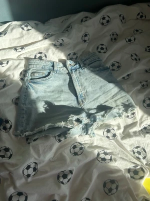 Ljusblå högmidjade jeansshorts - Säljer ett par ljusblå jeansshorts med hög midja och råa, fransiga kanter. Klassisk femficksmodell med knapp och dragkedja framtill. Perfekta för varma dagar och enkel att matcha med olika toppar.