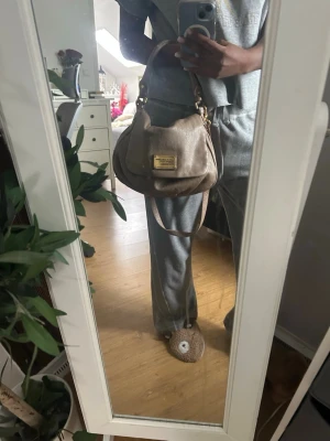 Beige axelremsväska i skinn - Snygg beige handväska i mjukt skinn med guldfärgade metalldetaljer och en tydlig logoplatta framtill. Väskan har både kort handtag och en längre axelrem så du kan bära den på olika sätt. Perfekt storlek för det viktigaste och ger en clean look till din outfit. Har slitage som stan i sista bilderna på handtaget och i tyget över loggan.