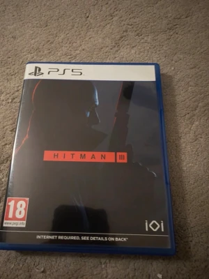 Hitman III till PS5 - Hitman III till PlayStation 5 i blå plastfodral. Omslaget är svart med en siluett av Agent 47 och röd text. Spelet är PEGI 18 och kräver internetanslutning. Perfekt för dig som gillar action och stealth-spel.