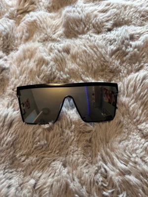 Svarta futuristiska solglasögon - Säljer ett par svarta solglasögon med spegelglas och futuristisk, kantig design. Glasögonen har en bred, rak båge och stora linser som täcker mycket av ansiktet. Perfekta för dig som vill sticka ut med en modern och edgy stil.