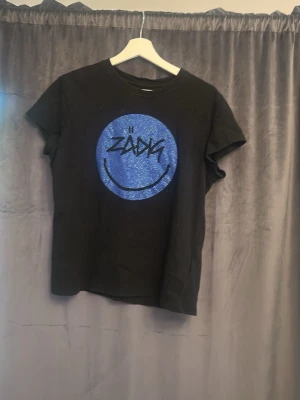 Svart Zadig & Voltaire t-shirt - Cool svart t-shirt från Zadig & Voltaire med ett stort blått smileytryck och texten 'ZADIG' på bröstet. Klassisk rund hals och korta ärmar. Perfekt för dig som gillar streetstyle och vill sticka ut lite extra.