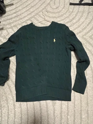 Mörkgrön kabelstickad tröja Polo - Mörkgrön kabelstickad tröja från Polo Ralph Lauren med klassisk gul broderad logga på bröstet. Tröjan har rund halsringning, ribbstickade muddar och nederkant. Perfekt för en stilren och avslappnad look.