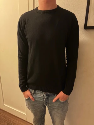 Svart basic rundhalsad tröja - En enkel svart långärmad tröja med rund halsringning. Tröjan har en rak passform och ribbade muddar vid ärmslut och nederkant. Perfekt att matcha med jeans eller under en jacka. Materialet ser ut att vara mjukt och stretchigt.