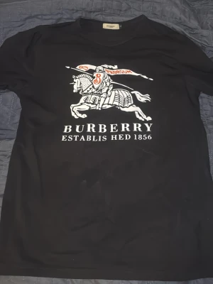 Svart Burberry t-shirt med tryck - Svart t-shirt med stort Burberry-tryck framtill som visar en riddare på häst och texten 'BURBERRY ESTABLISHED 1856'. Klassisk rund halsringning och korta ärmar. Perfekt för dig som gillar statement-plagg.