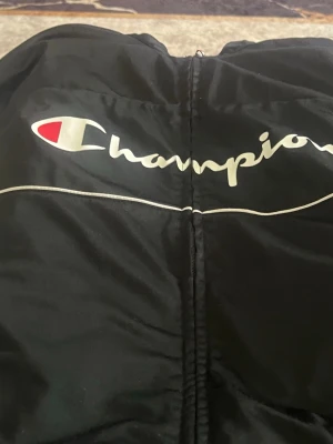 Svart zip-jacka från Champion - Säljer en svart zip-jacka från Champion med stor logga över bröstet i vitt och rött. Jackan har en sportig look med vita detaljer och är tillverkad i ett glansigt syntetmaterial. Perfekt för dig som gillar streetwear och vill ha en stilren jacka.