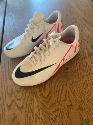 Vita Nike fotbollsskor med neonröda detaljer - Säljer ett par vita Nike fotbollsskor med svart swoosh och neonröda grafiska detaljer på sidorna. Skorna har snörning och låg profil, perfekta för snabba rörelser på planen. Tillverkade i syntetmaterial för bra grepp och känsla.perfekt som inomhus sko till idrotten. Sparsamt använda storlek 38 ord pris 800kr mitt pris 500kr 