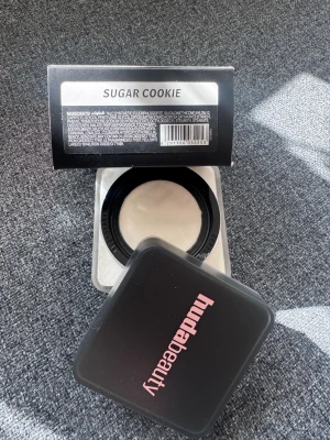Huda Beauty Sugar Cookie puder - Transparant löspuder i nyansen Sugar Cookie från Huda Beauty. Kommer i en fyrkantig burk med svart lock och rosa logga. Perfekt för att fixera foundation och ge en matt finish. Pudret är vitt och har en silkeslen textur.