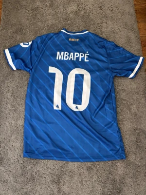 Blå Real Madrid matchtröja Mbappé - Snygg blå Real Madrid matchtröja med vita detaljer och tryck. Mbappé och nummer 10 på ryggen, samt klubbmärke och Adidas-logga på bröstet. Tillverkad i lätt och ventilerande material, perfekt för fotboll eller träning.