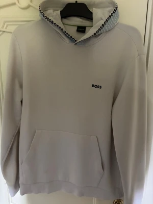 Vit hoodie från BOSS - Snygg vit hoodie från BOSS med diskret logga broderad på bröstet och BOSS-tryck längs insidan av huvan. Klassisk känguruficka framtill och ribbade muddar. Perfekt för en clean och stilren look.