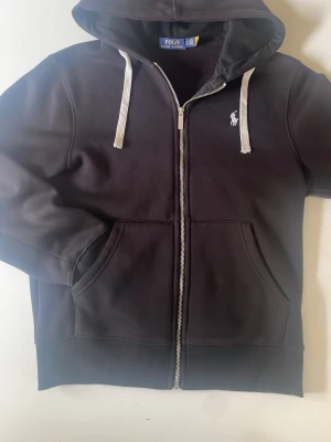 Svart hoodie från Polo Ralph Lauren - Svart hoodie med dragkedja från Polo Ralph Lauren. Klassisk design med vit logga på bröstet, stora fickor fram och vita snören i huvan. Tillverkad i mjukt material och har ribbade muddar vid ärmslut och nederkant. Perfekt för en avslappnad stil.