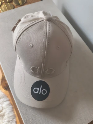 Beige keps från Alo - Snygg beige keps från Alo med broderad logga framtill och justerbar rem bak. Klassisk böjd skärm och ventilerande hål upptill. Tillverkad i mjukt bomullstyg som ger en clean och stilren look.