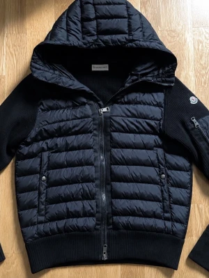Svart pufferjacka med huva  - Snygg svart pufferjacka  med quiltad front och huva. Jackan har ribbstickade ärmar och sidor, två fickor med tryckknappar och dragkedja. Snygg svart cardigan oerfekt till höst och vinter, i strolek L men passar även M och S